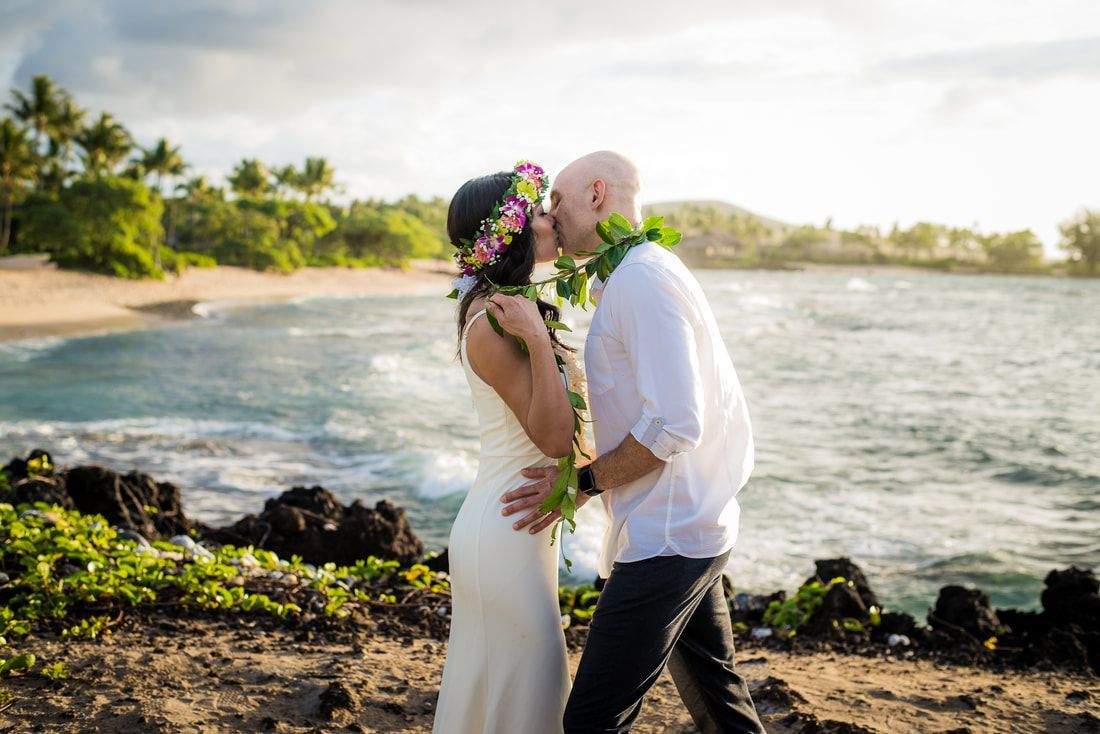 A Simple Kona Beach Weddings couple kissing on a beach. Bride wears a floral crown, groom has a lei. Ocean and palm trees in background.