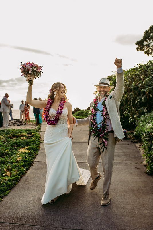 Newlyweds celebrate, raising arms. Woman wears white gown, man a suit, both with leis, on a path.