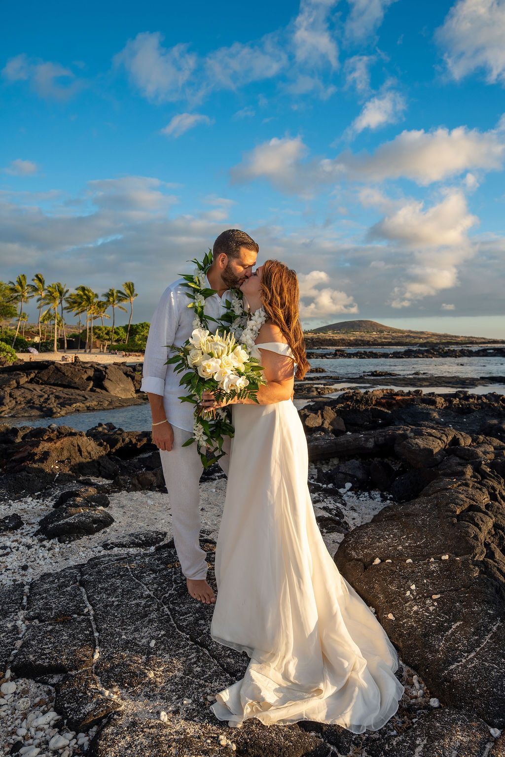 Couple kissing on rocky shore at sunset; bride holds bouquet, groom wears lei.