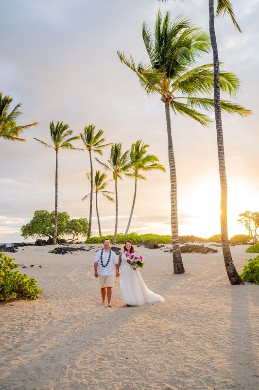 Bride and groom hold hands on beach, palm trees, sunset.
