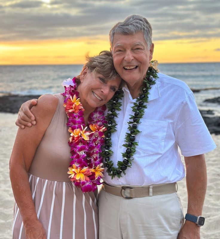 Couple on beach, embracing, wearing leis, smiling at sunset.