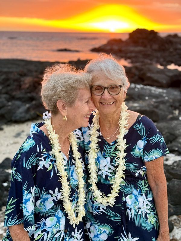 Two women in matching floral shirts and leis, smiling, embracing at sunset on a rocky beach.