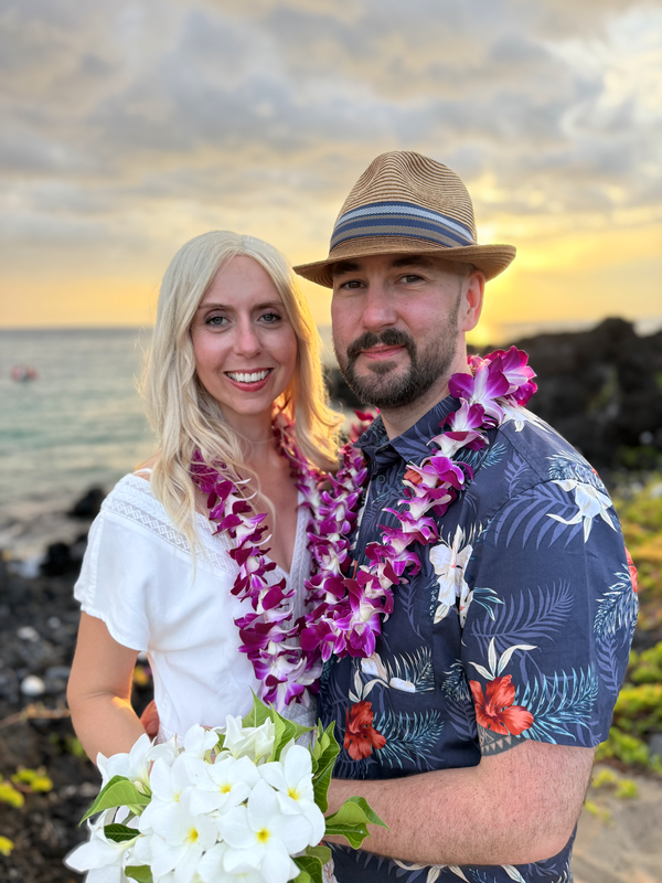 Couple wearing leis on beach at sunset. Woman smiles, holds flowers; man in hat smiles.