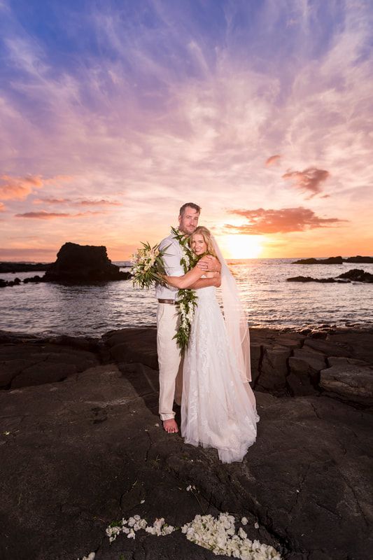 Couple embraces on rocky beach at sunset. Bride in white dress, groom in tan pants and shirt. Ocean and colorful sky.