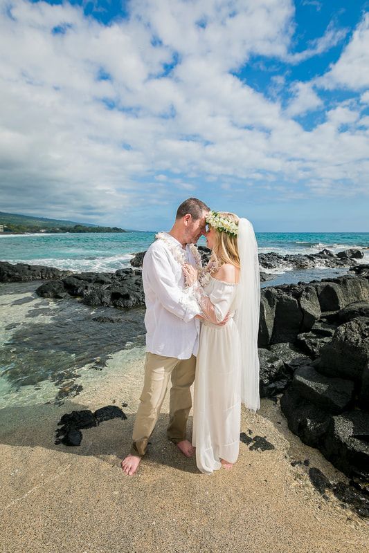 Couple embraces on a beach, woman in wedding dress and floral crown, man in white shirt and tan pants.