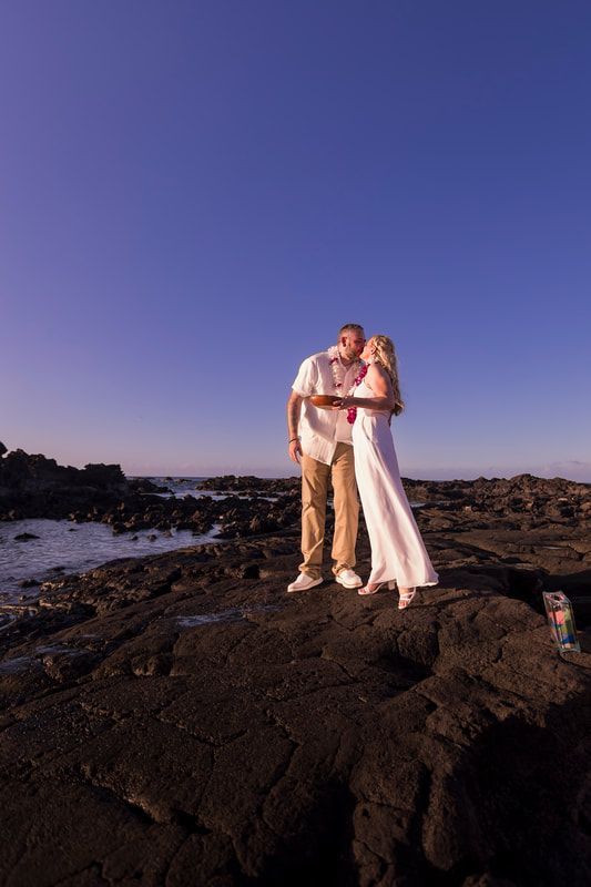 Couple kissing on black rocks by the ocean at sunset; the woman wears a white dress.