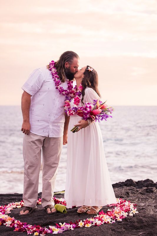 Couple kissing on a beach, surrounded by flower petals. The sky is pink, and the ocean is visible.