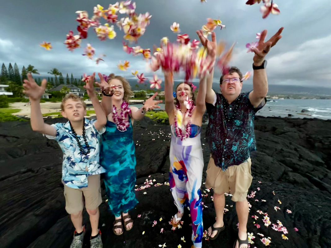 Family throwing flower petals into the air on a rocky coast; cloudy sky.