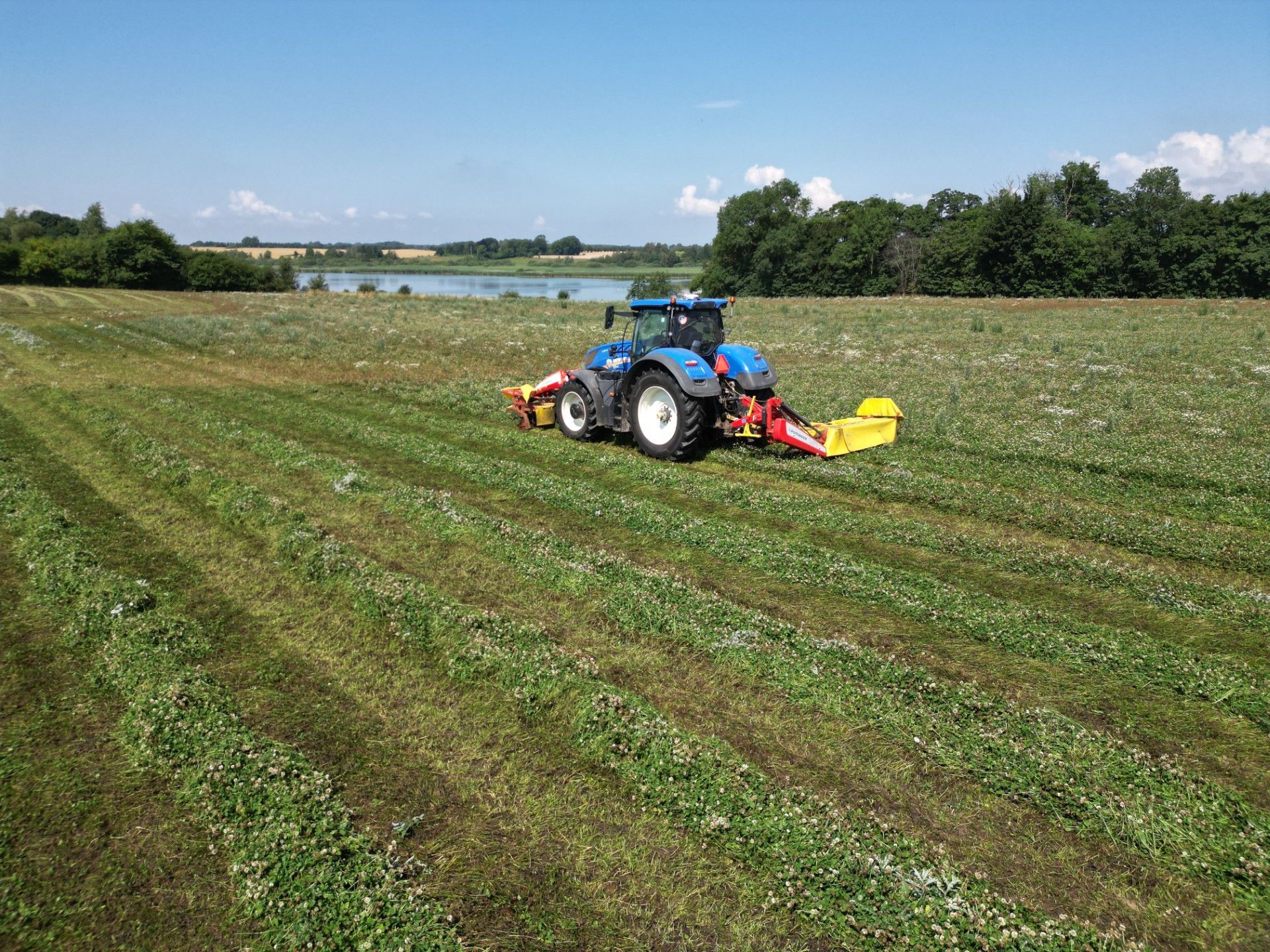 Blå traktor slår hø i en grøn mark under en delvist overskyet himmel med en sø og træer i det fjerne