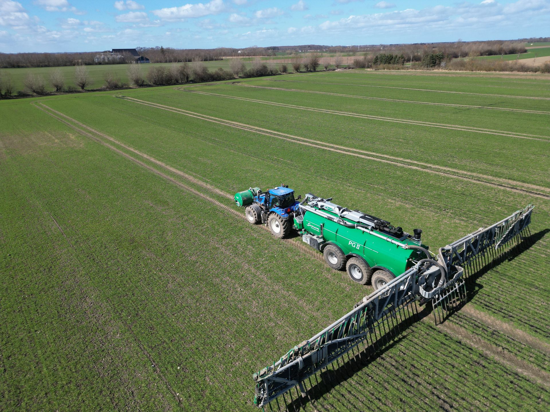 Blå traktor trækker en grøn sprøjte hen over en stor mark under en klar himmel