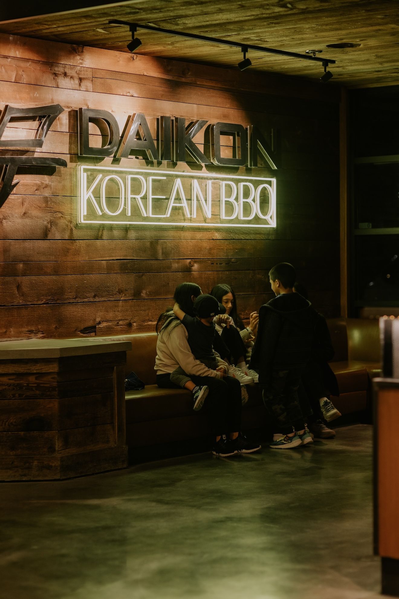 a group of people are sitting on a couch in front of a neon sign that says daikon korean bbq .