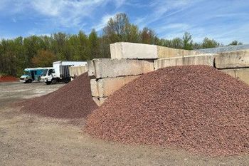 Outdoor gravel yard with large reddish-brown stone piles, concrete blocks, and trucks in the background