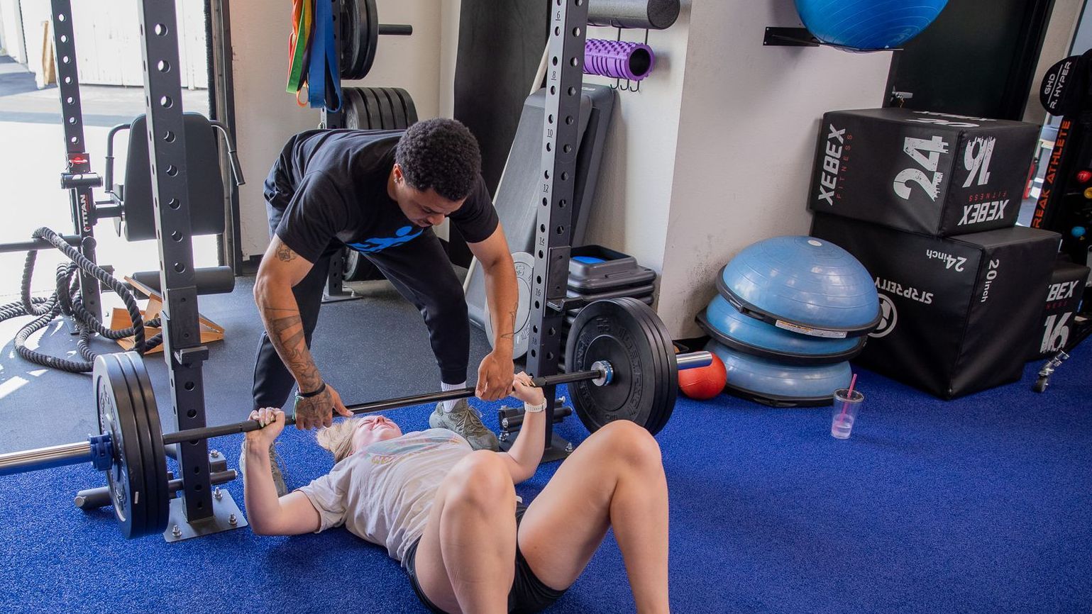A man is helping a woman stretch her legs in a gym.
