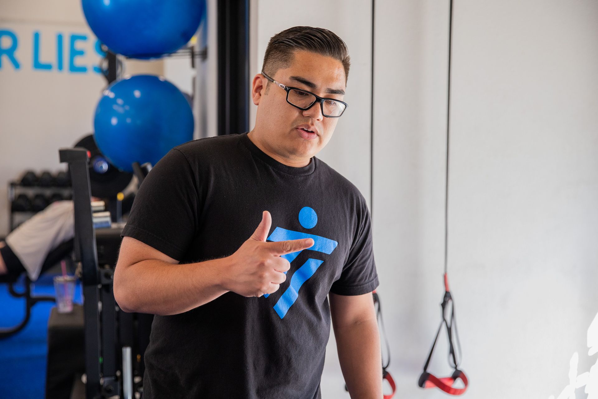 Man in glasses points to the side while speaking in a gym.