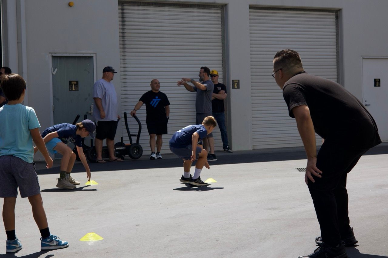 Children participating in an outdoor sports drill, overseen by an instructor, with spectators in the background.