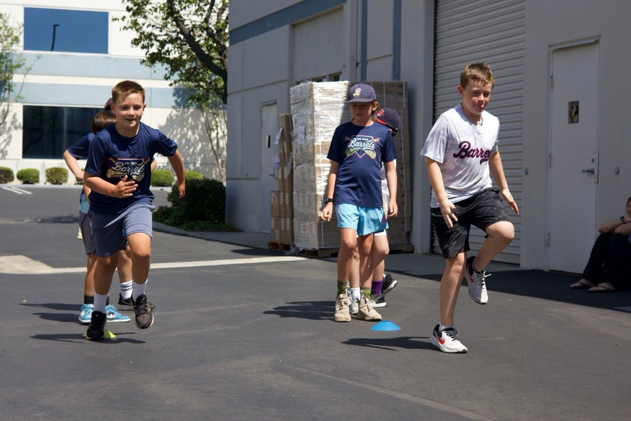 Children run across an outdoor parking lot during an athletic activity near building walls.