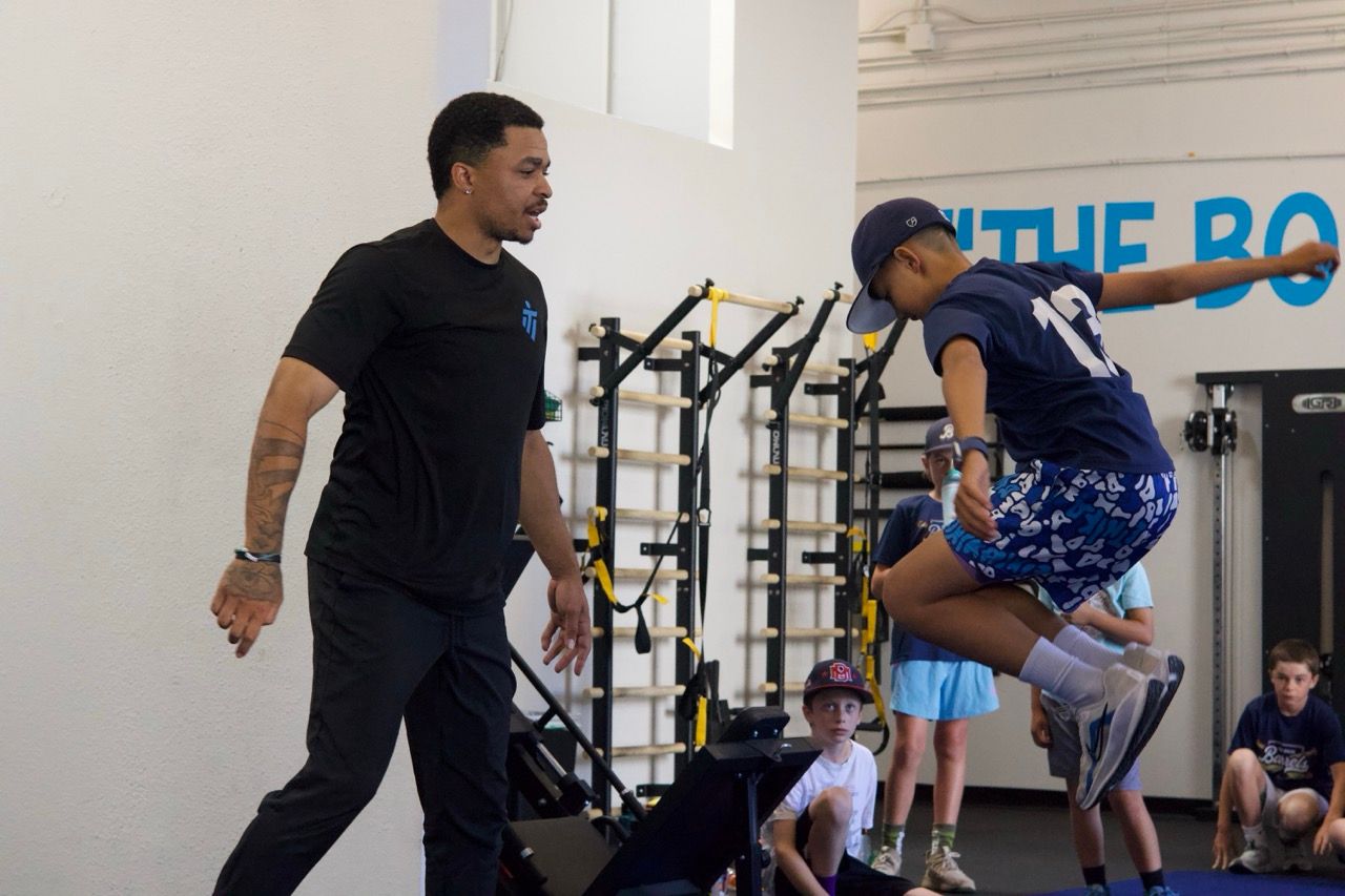 A coach watches as an athlete performs a jump in a gym, with other people observing in the background.
