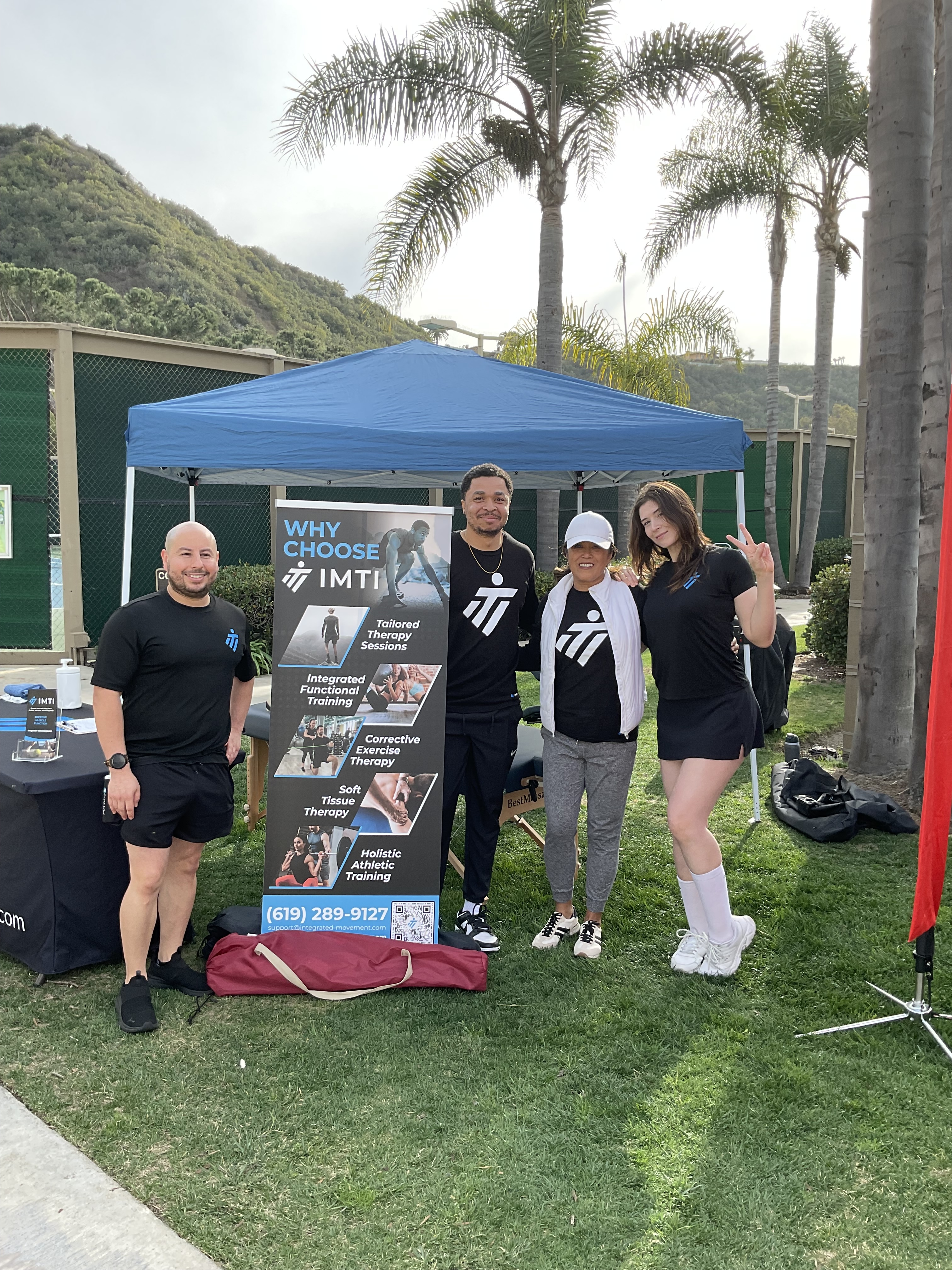 Group of four people standing in front of a booth with a banner, outdoors. They wear black attire with a logo; background includes greenery and a blue canopy.