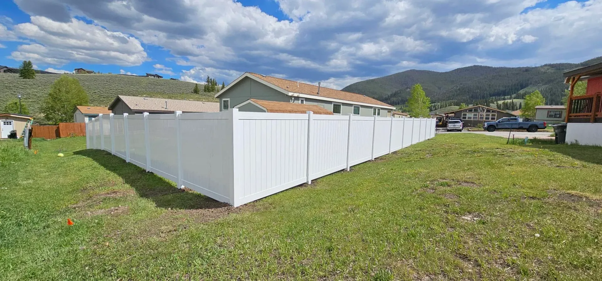 White vinyl fence around a grassy yard under a partly cloudy sky, with houses and mountains in the background