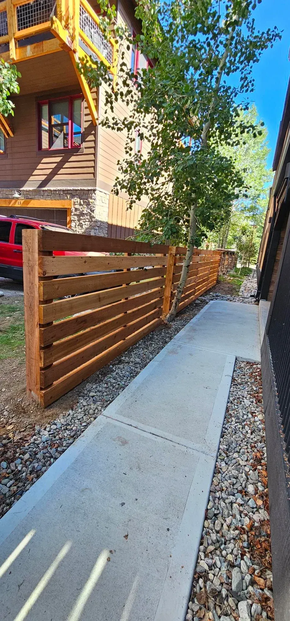Wooden fence beside a narrow concrete walkway next to a building with trees and gravel landscaping.