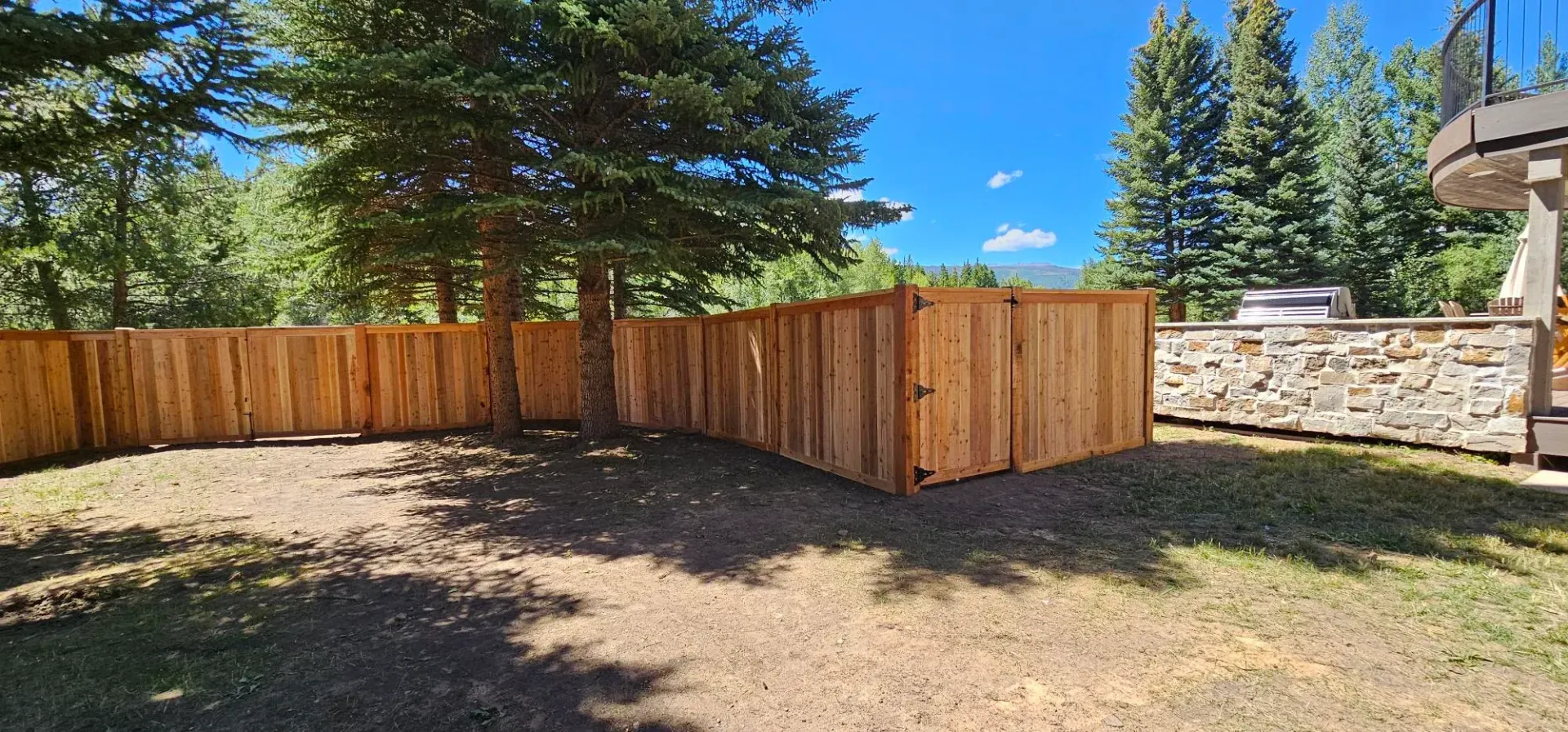 Wooden privacy fence and gate in a sunny backyard with trees and stone wall