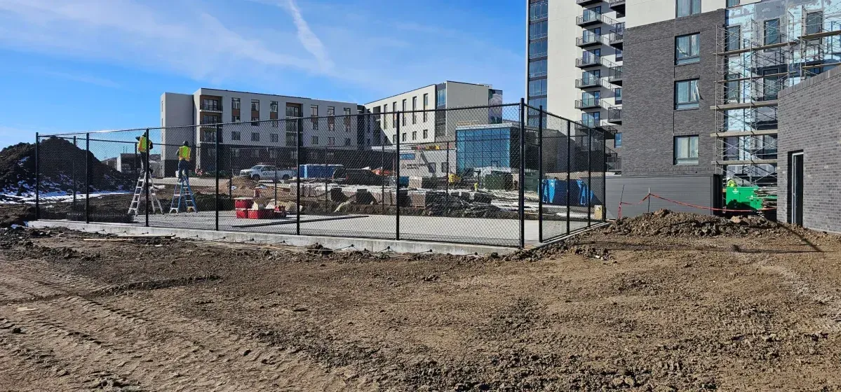 Construction site with dirt foreground, fenced area, and modern apartment buildings under a blue sky