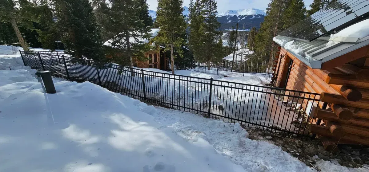 Snowy yard with black fence, pine trees, and a cabin beside a mountain view