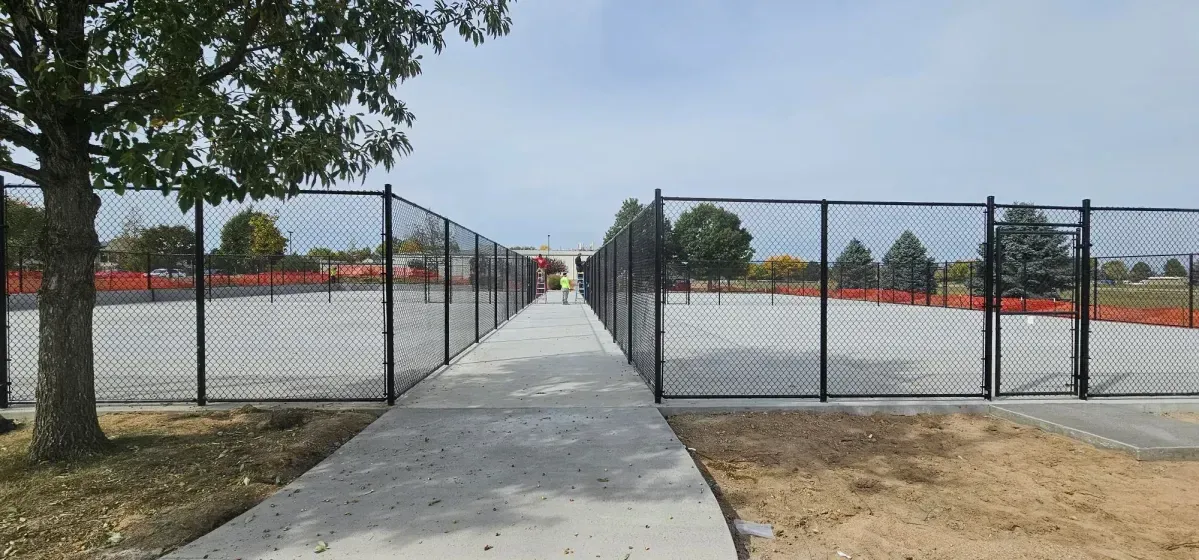Concrete walkway leading through a black chain-link gate into a fenced outdoor area with bleachers on both sides