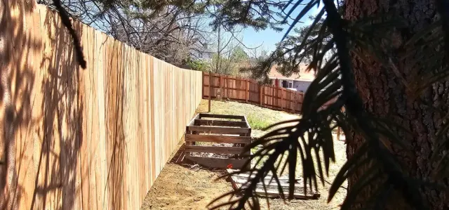 Narrow backyard path beside a wooden fence, with trees casting shadows and a brick wall at the end.