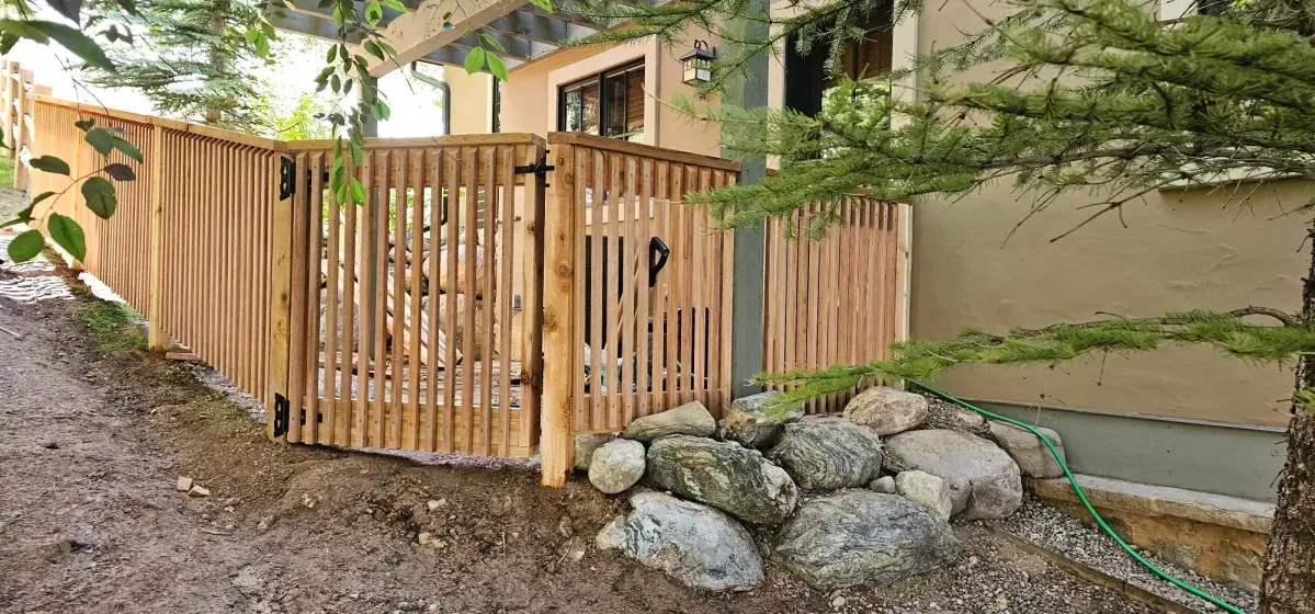 Wooden gate and fence around a yard beside a house, with rocks and trees in the foreground.