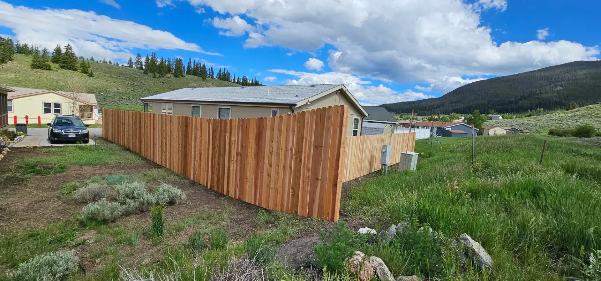 Wooden fence around a small building in a grassy rural lot with mountains and clouds in the background