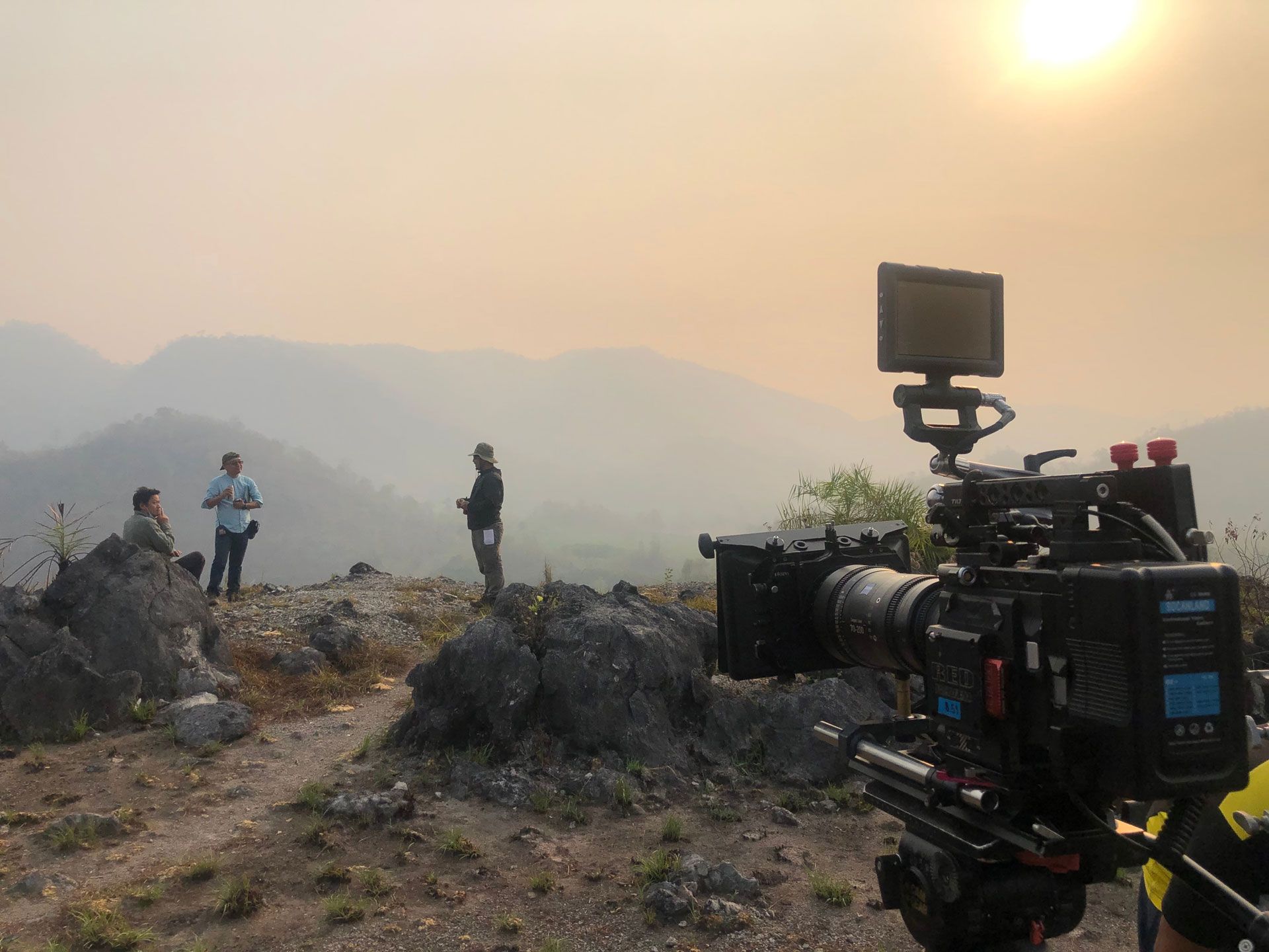 A professional video camera on a tripod faces three people standing on a rocky, hazy mountain ridge at sunset.
