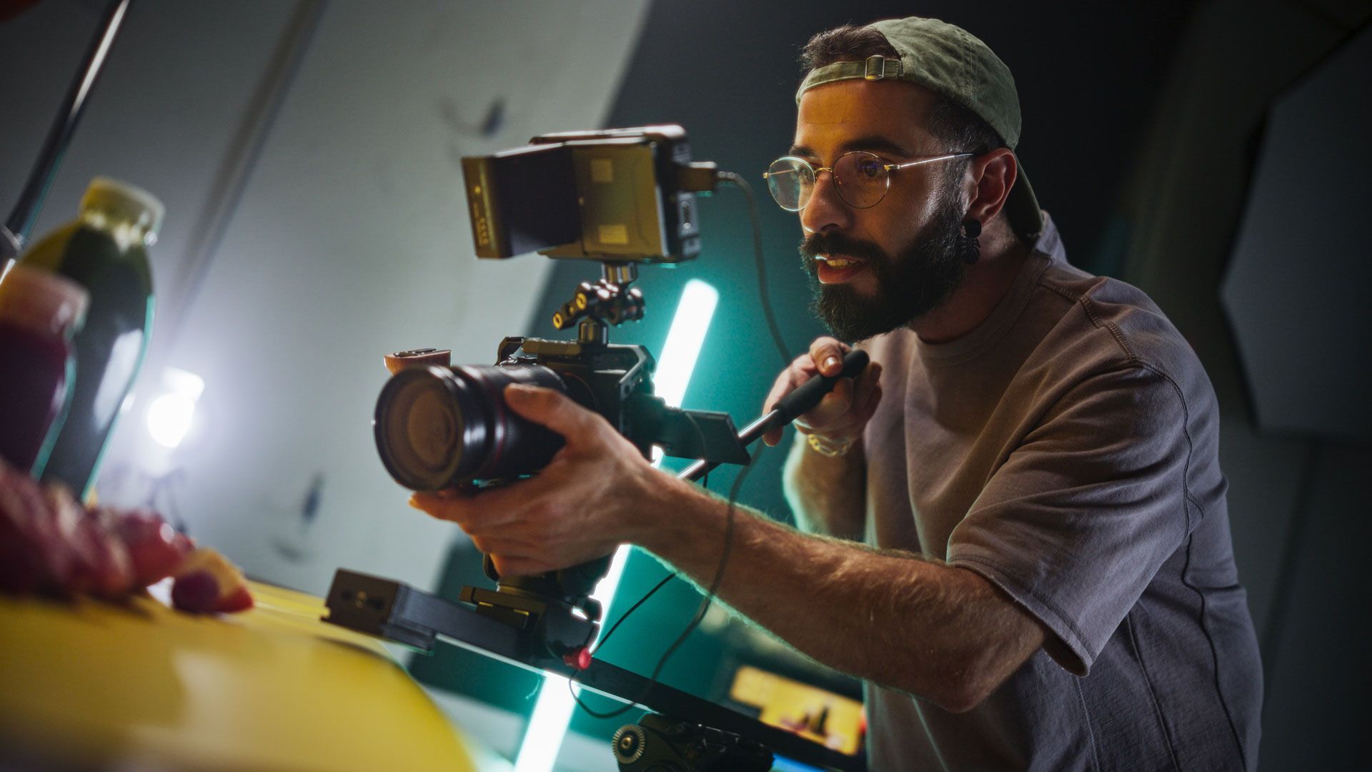 A person wearing a cap and glasses focuses a professional camera on food placed on a yellow table in a dark studio.