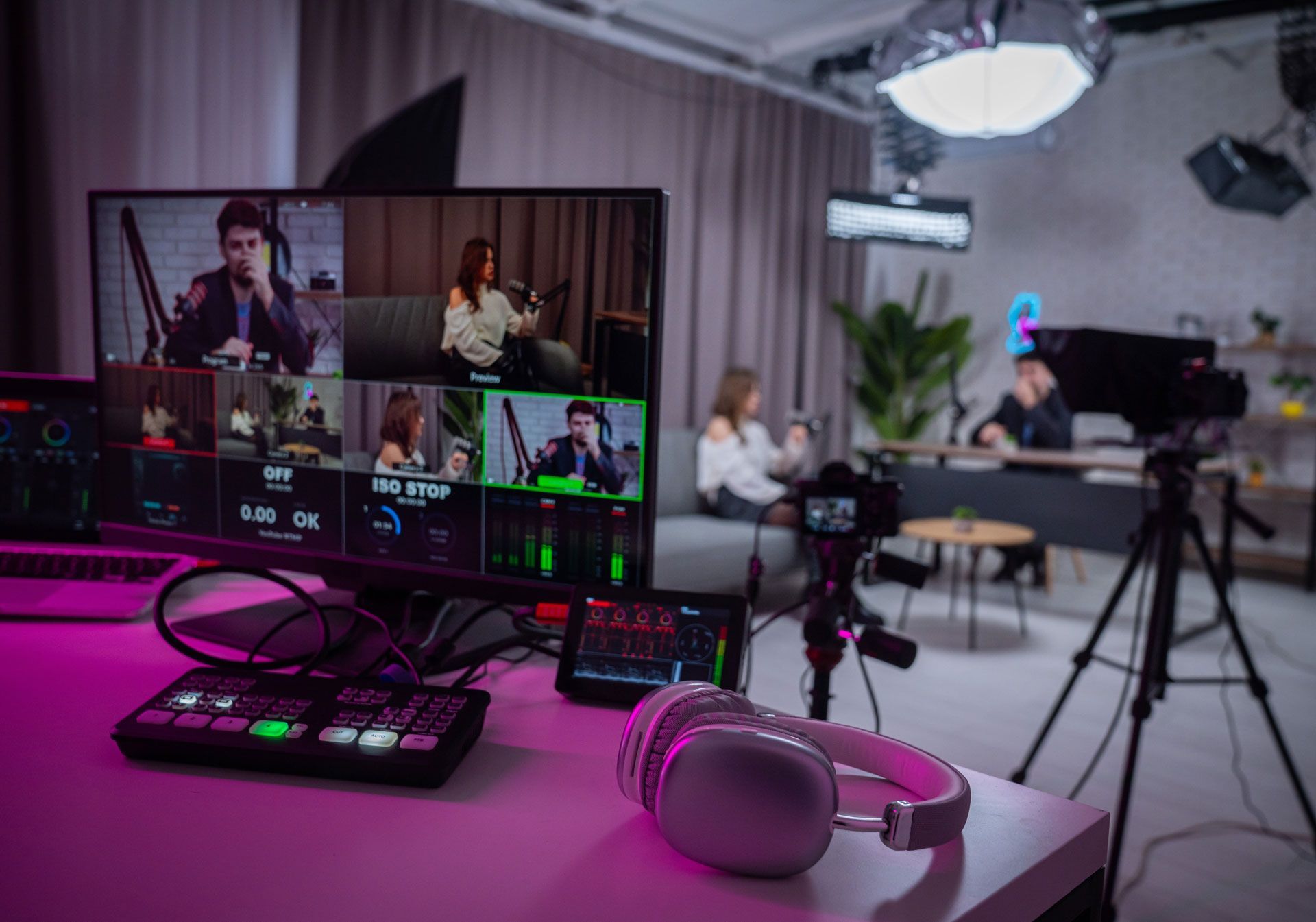A monitor showing a video production dashboard sits on a desk with headphones, overlooking a lit studio set.
