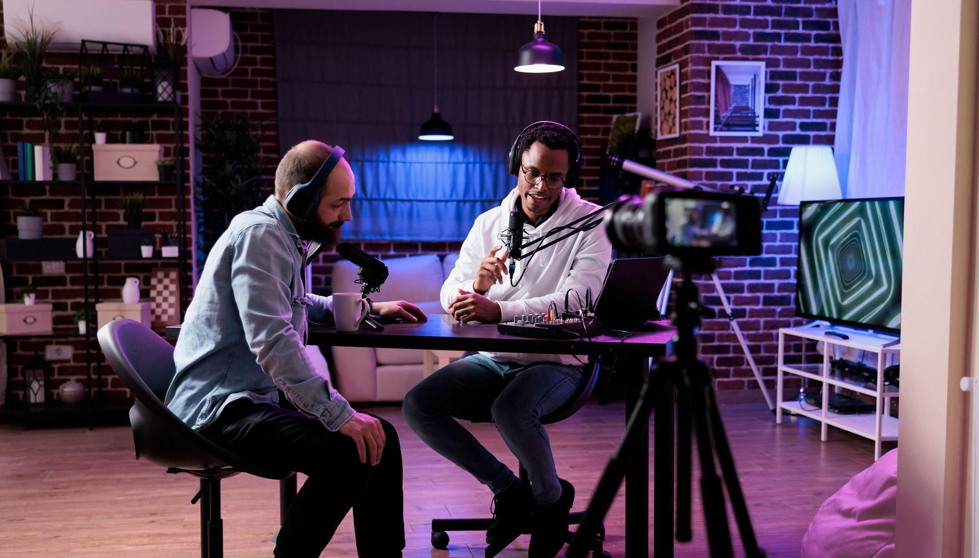 Two people recording a podcast at a desk in a home studio, with mics, camera, and ambient lighting.