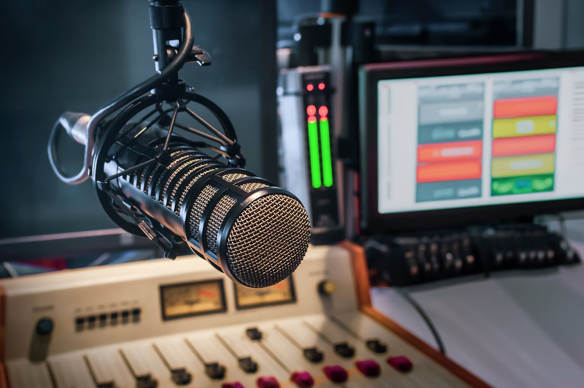 Close-up of a microphone in a radio studio. Soundboard and computer monitor with audio levels in the background.
