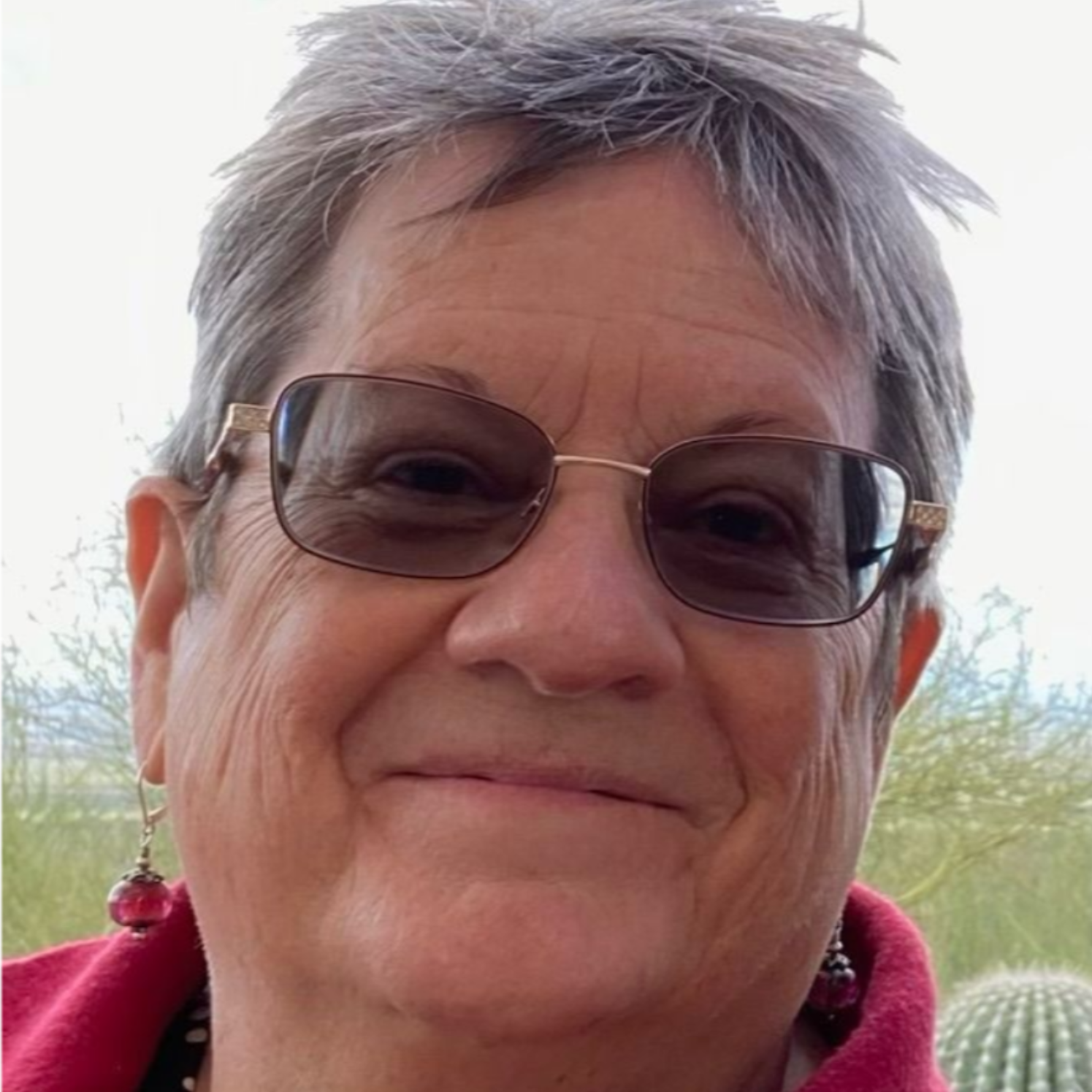 Smiling person wearing sunglasses and a red jacket outdoors with greenery in the background
