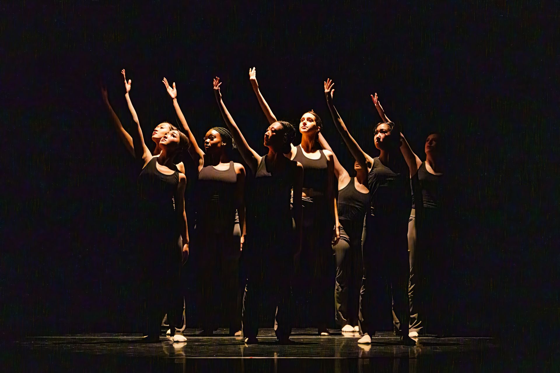 Five dancers in black costumes striking synchronized poses on a dark stage under warm spotlight.