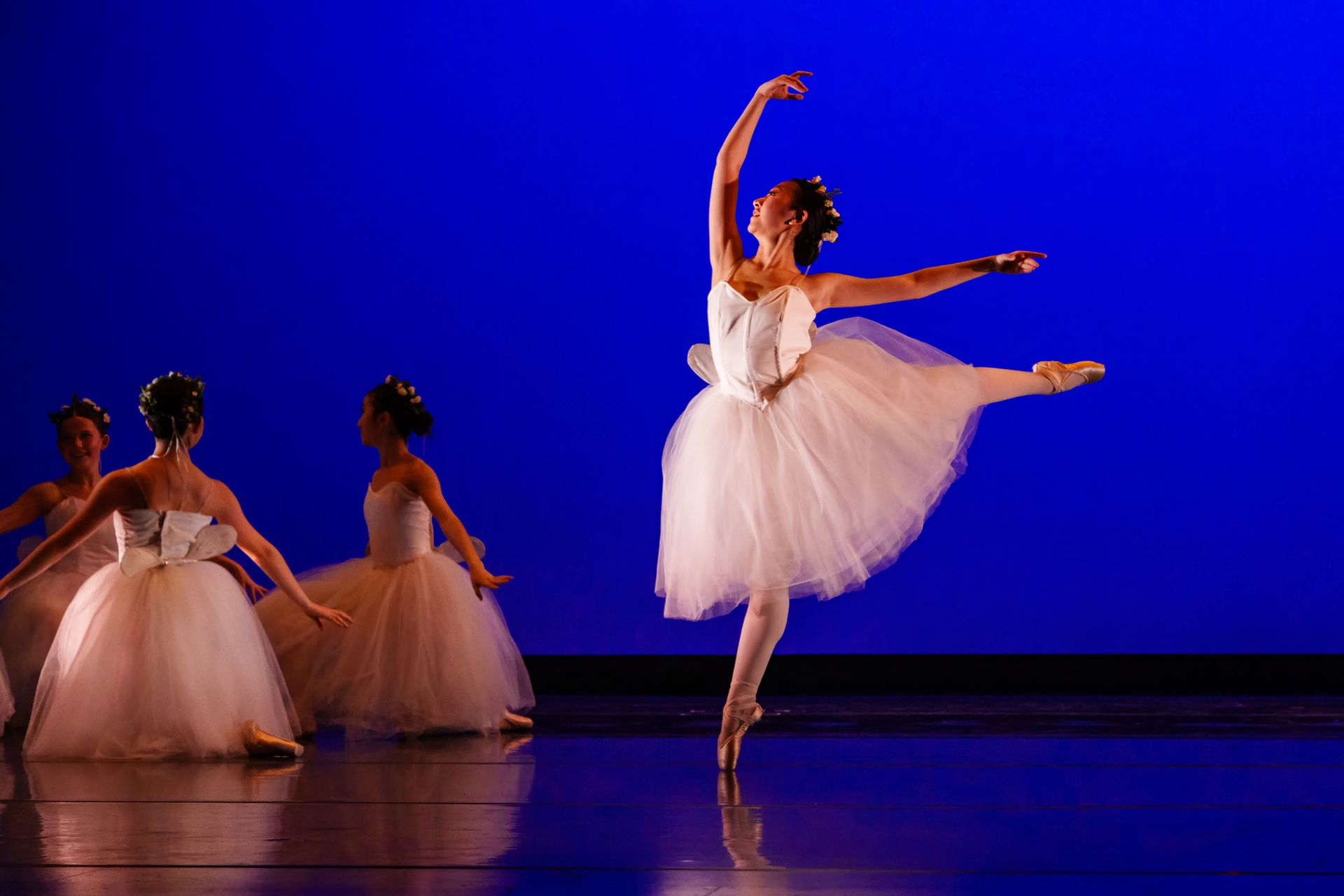 Ballet dancers in white tutus on a stage, one performing a high leg extension under blue lighting