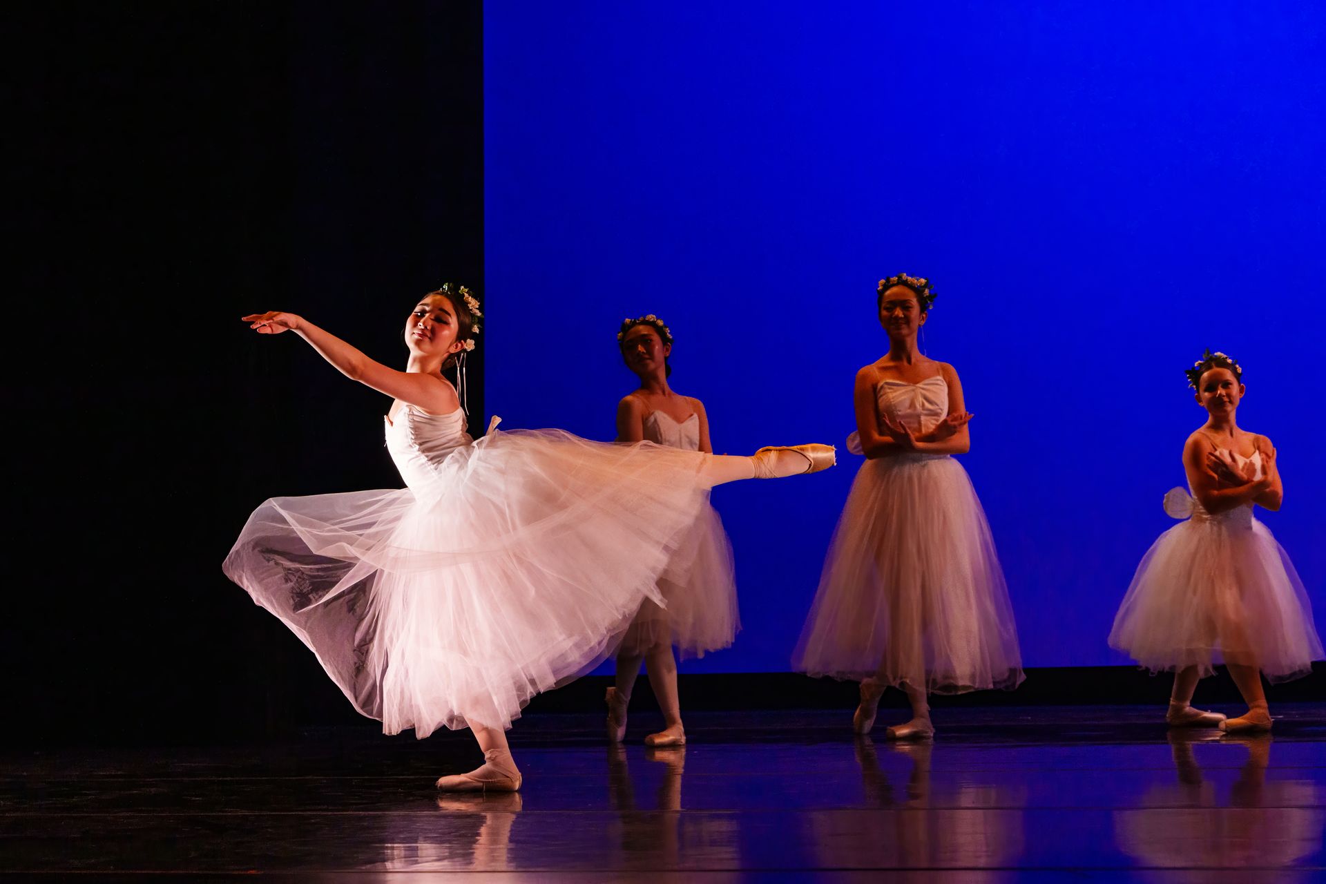 Ballet dancers onstage, one in a white tutu mid-leap before three dancers in pink costumes against a blue backdrop