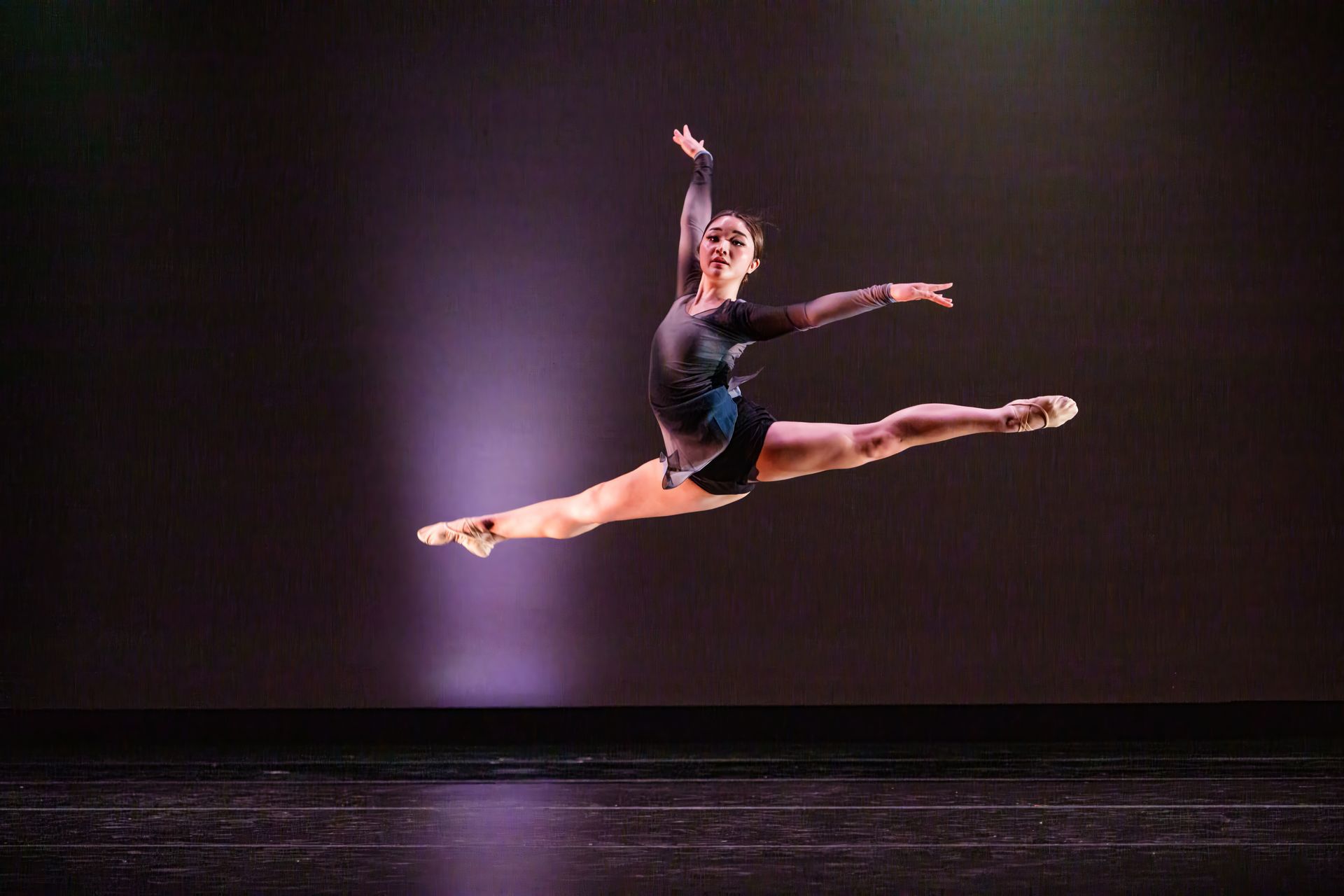 Ballet dancer midair in a split leap on a lit stage against a dark purple backdrop