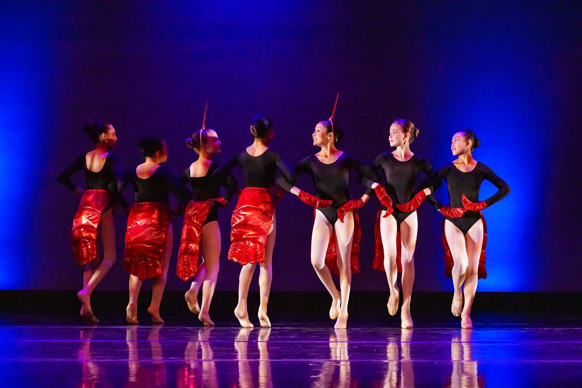 Six dancers in black and red costumes perform on a blue-lit stage