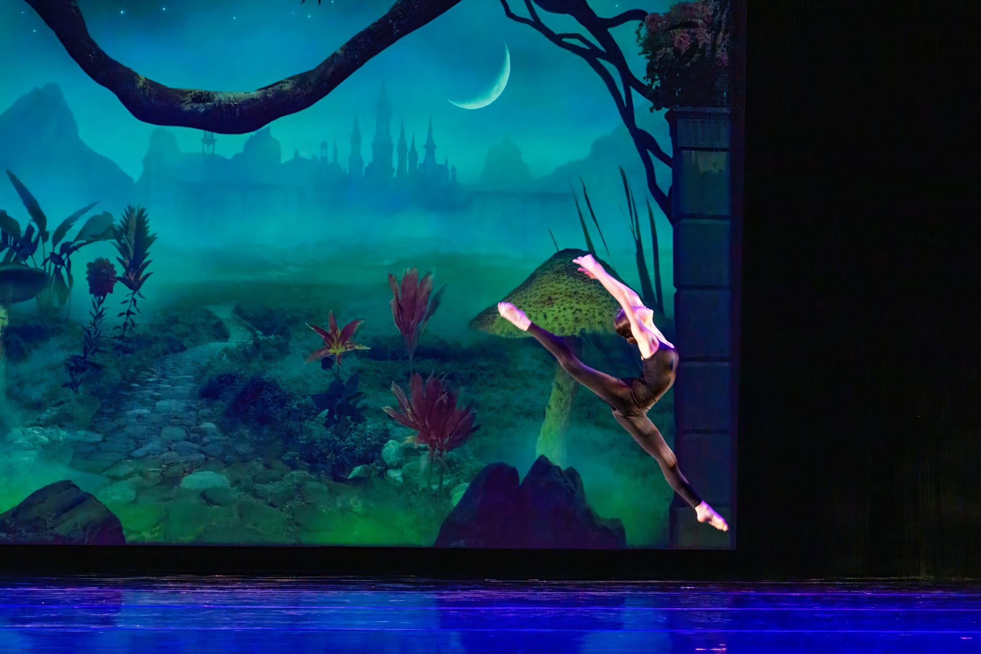 Dancer in white leaps against a blue-green jungle backdrop on a dimly lit stage
