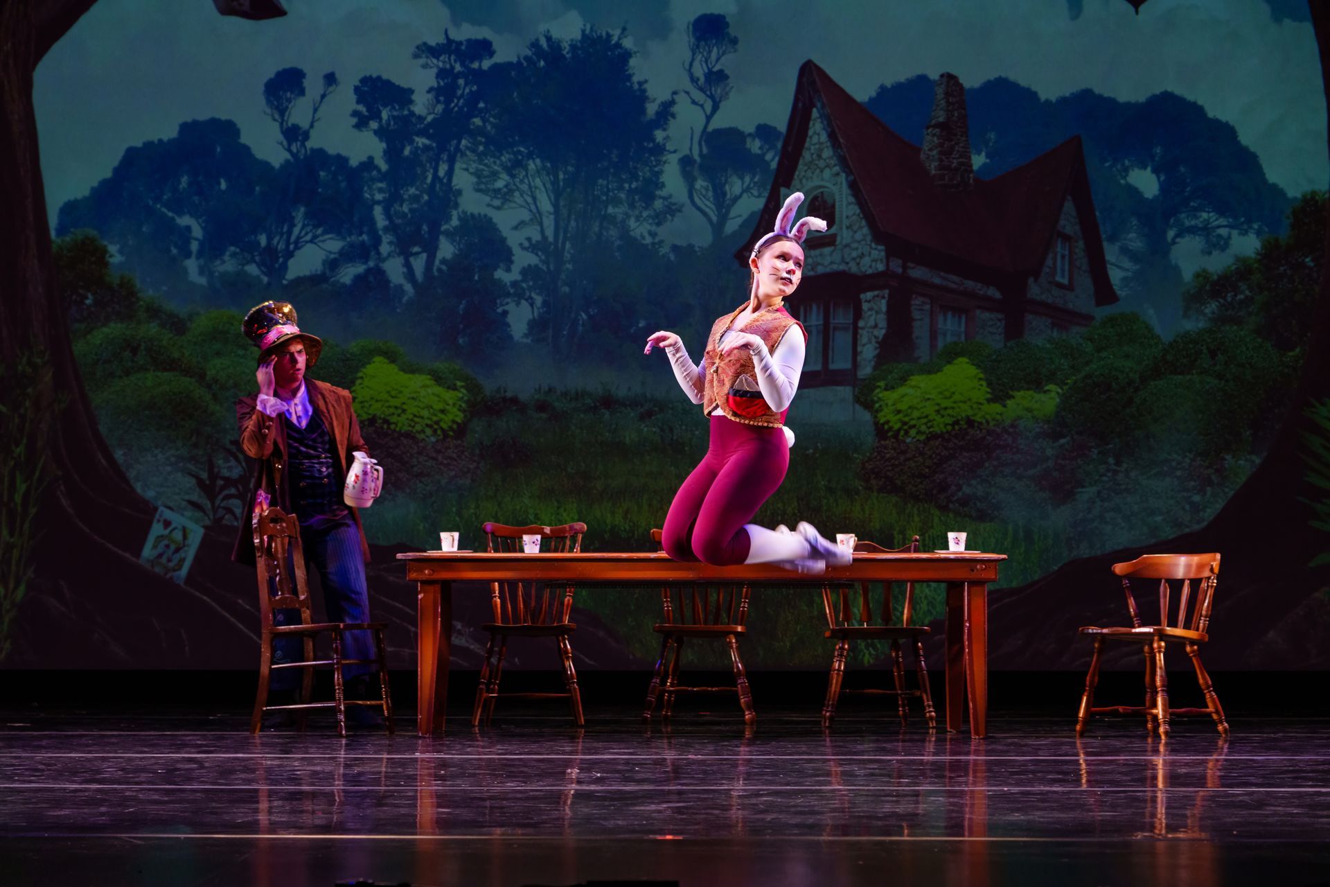 Two performers on a lit stage beside a long table, with a painted rural house backdrop at dusk.