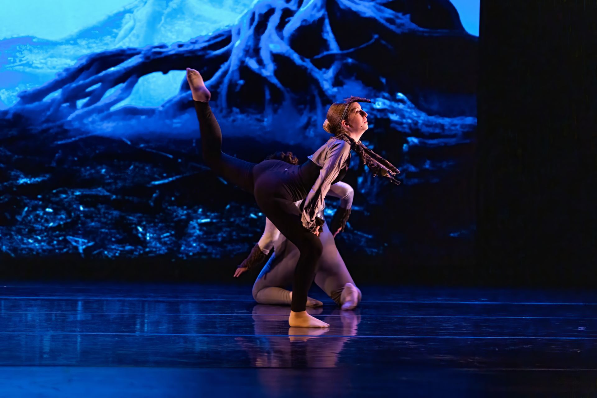 Two dancers in a dramatic blue-lit stage pose with raised limbs before a mountain backdrop