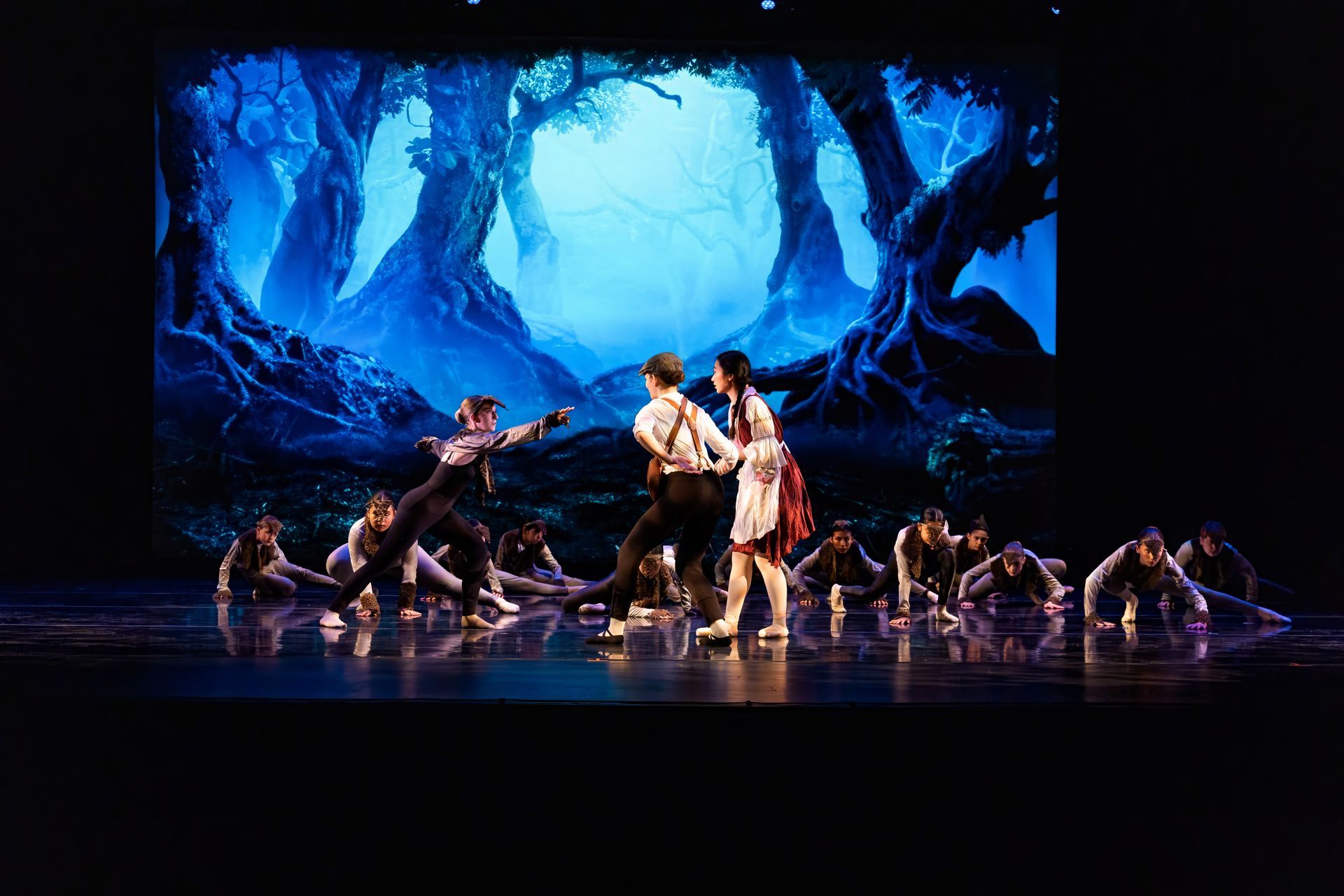 Dancers in white and dark costumes perform on a blue-lit stage with a forest backdrop and spiderlike figures.