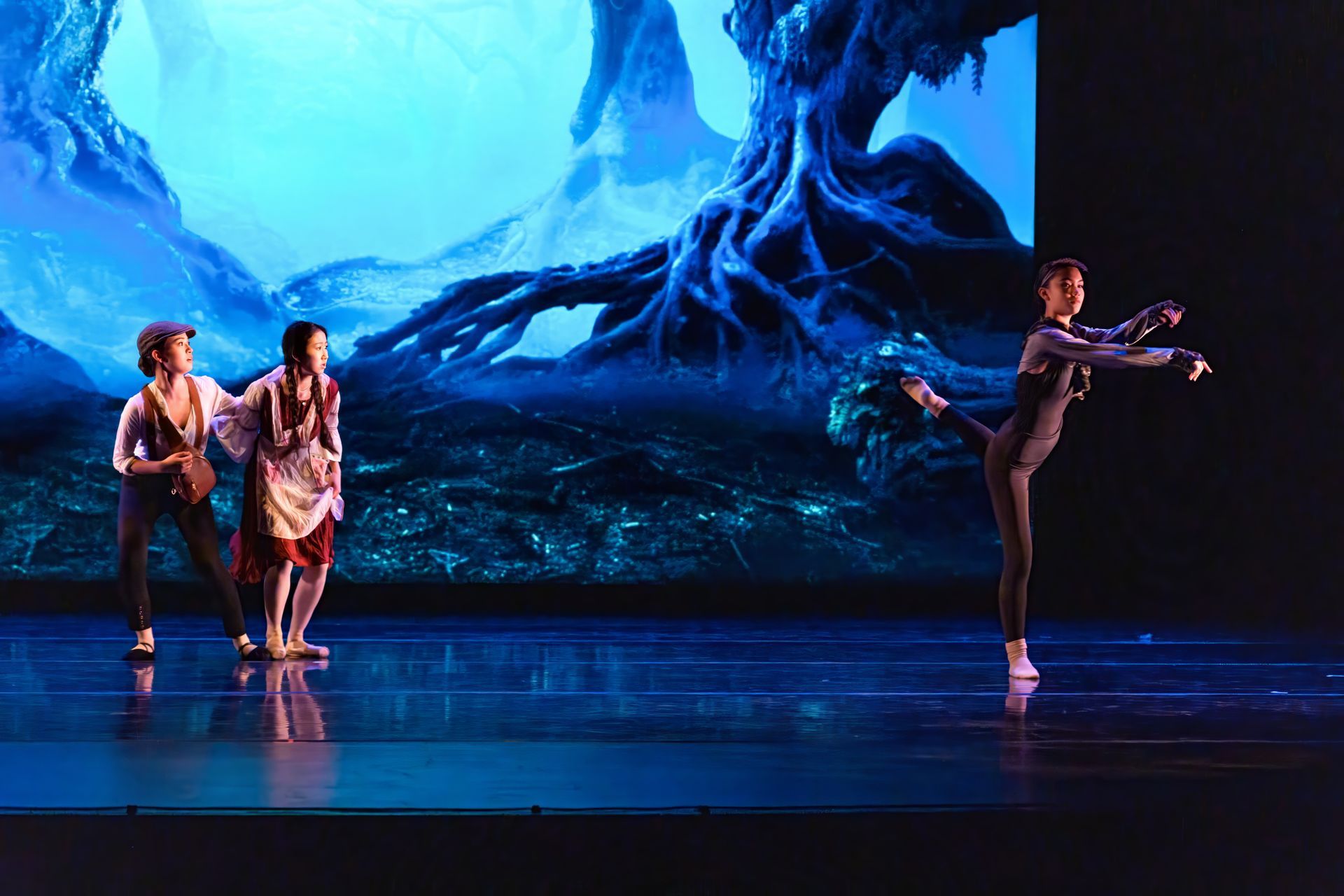 Three dancers on stage before a blue forest backdrop, one balancing in a high leg extension.