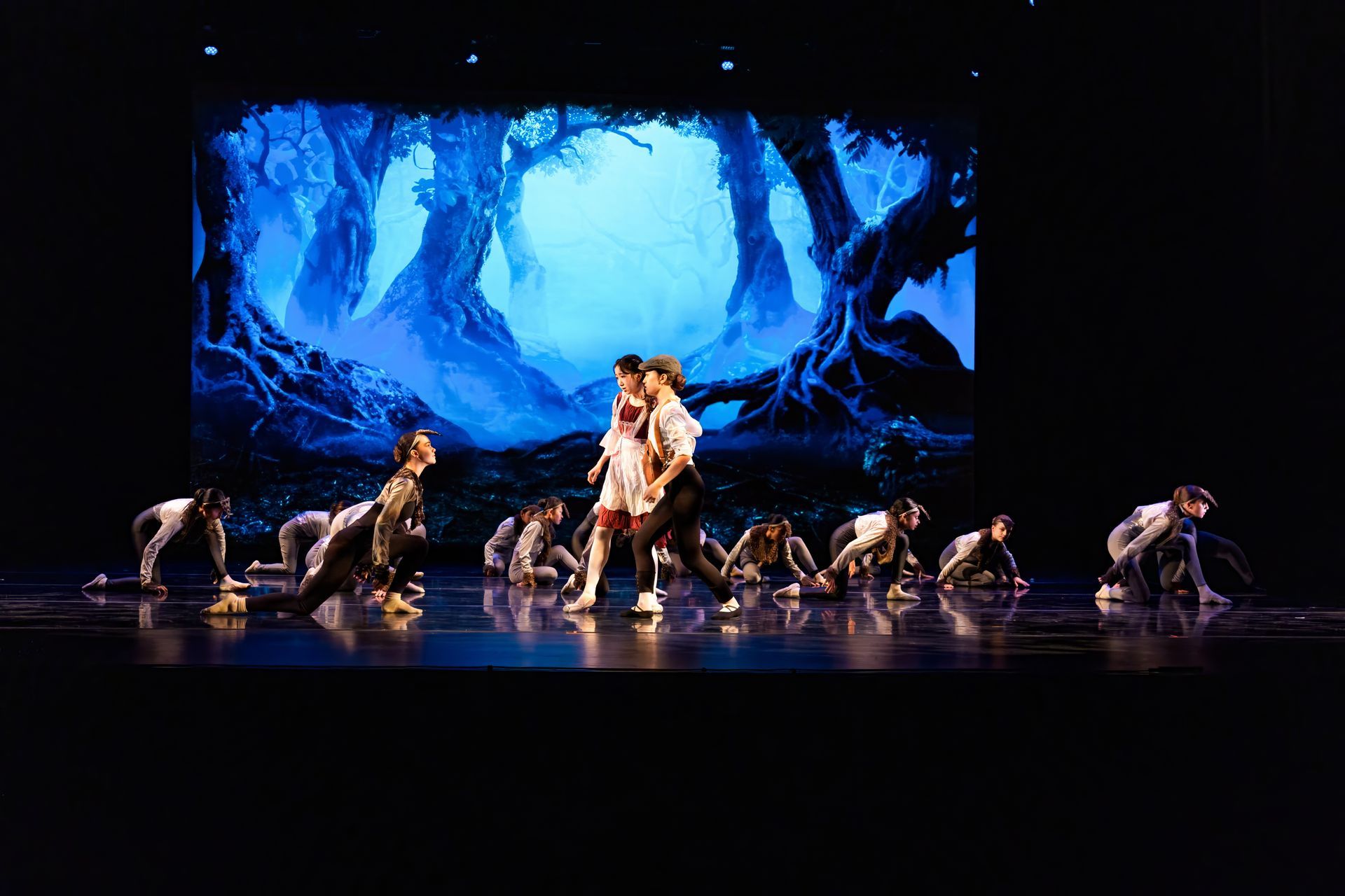 Dancers on a dark stage before a blue forest backdrop, with two central performers and others crouched around them