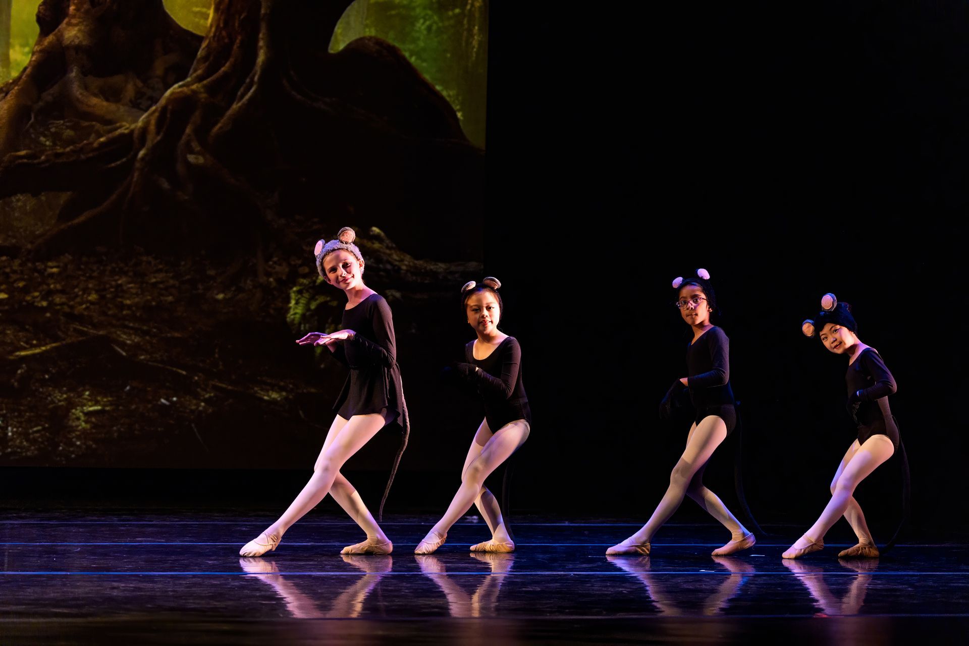 Four dancers in black costumes pose on a lit stage, with one extending a leg in front.