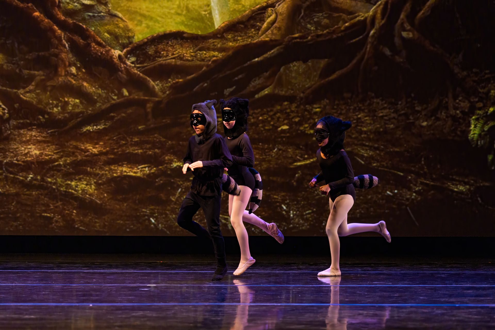 Three dancers in black costumes perform on a stage with a glowing forest backdrop.